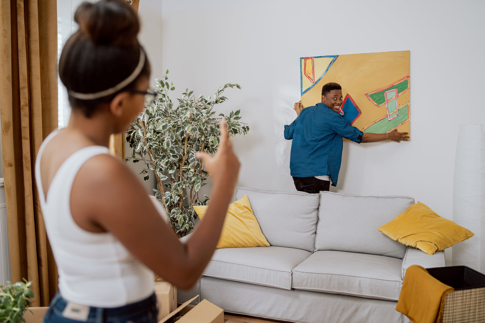Couple in love decorating their first apartment, unpacking boxes after moving in, the man hanging a painting on the wall, the woman showing how much to lift up