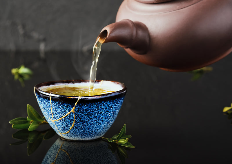 Hot green tea is poured from a ceramic teapot into a bowl. Selective focus on the blue cup. Steam will rise above the mug Reclaimed ceramic blue cup, second life of things, recycling or kintsugi