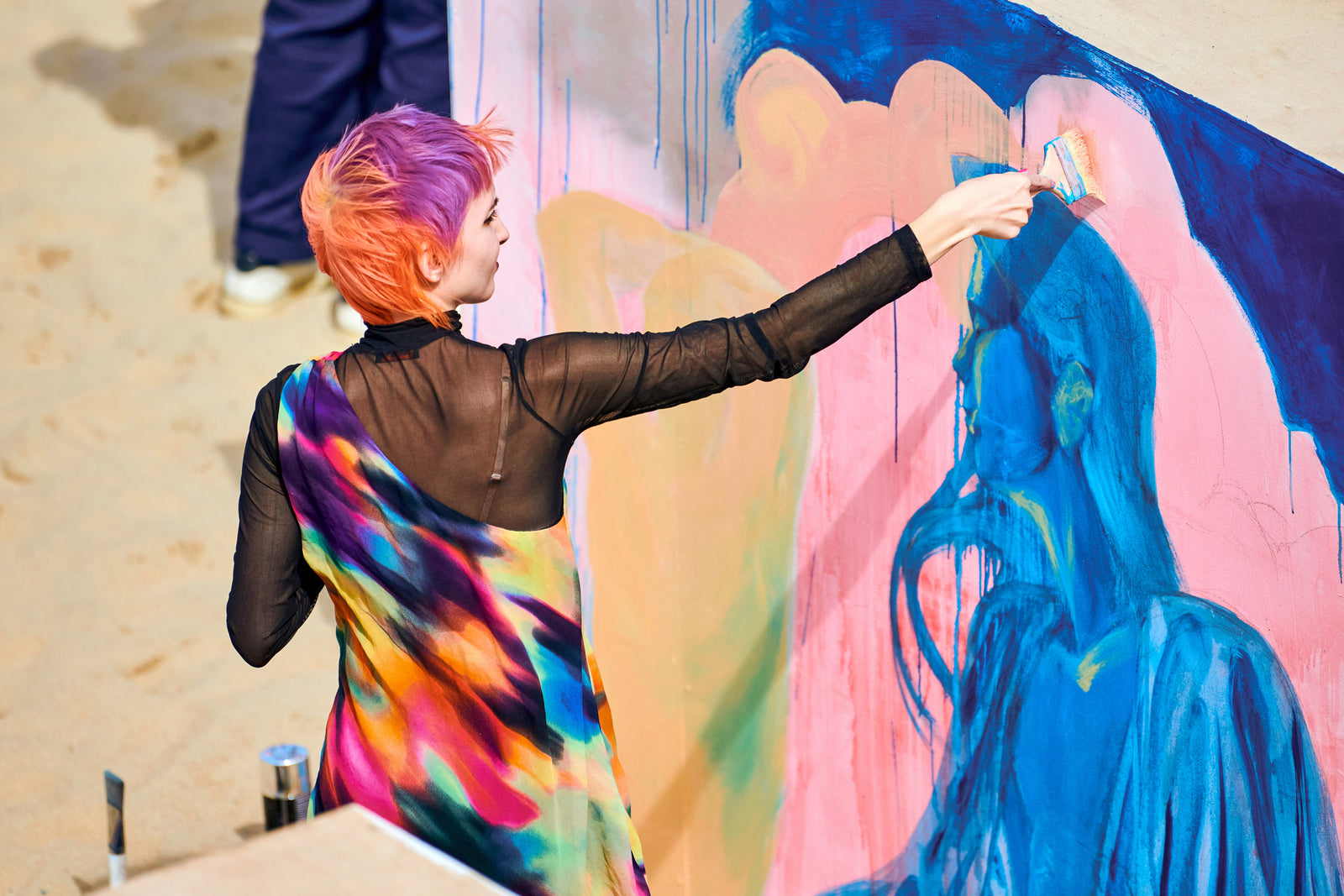 Young woman artist in bright colorful clothes painting multicolored picture on large canvas outside