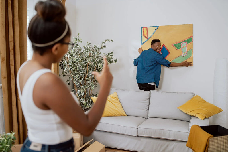 Couple in love decorating their first apartment, unpacking boxes after moving in, the man hanging a painting on the wall, the woman showing how much to lift up